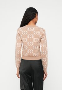 Sweater castanho claro com padrão de logótipo branco, mangas e bainha caneladas. Design justo, tecido de malha texturizado. Vista traseira, usado com calças pretas.