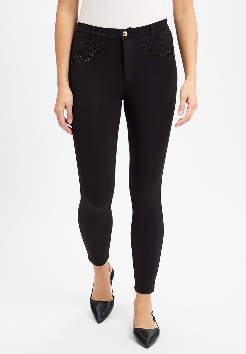 Pantaloni neri skinny con un design aderente, caratterizzati da dettagli con borchie nelle tasche anteriori e una chiusura con bottone dorato. Texture liscia.