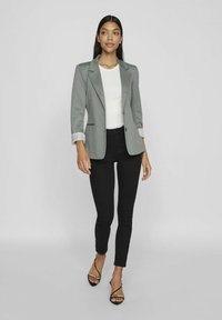 Grauer Blazer mit strukturiertem Design und gestreiften Ärmeln, getragen über einem weißen Top, kombiniert mit schwarzen Skinny-Jeans und schwarzen Riemchenschuhen.