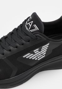 Zapatilla deportiva negra con parte superior de tela texturizada, cordones planos negros y detalle de logo plateado con alas en el lateral. Suela de goma con diseño antideslizante.