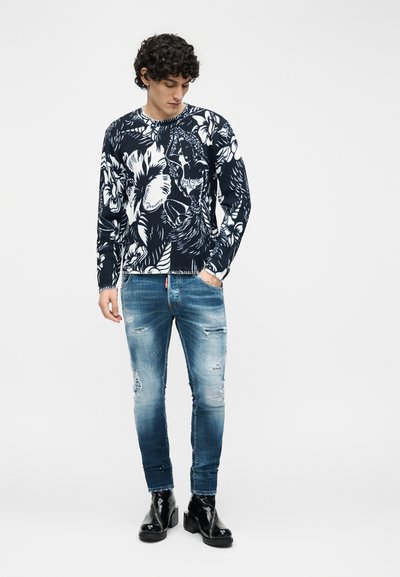 Homme portant un pull sombre à motif floral, un jean bleu déchiré et des bottes à talons noires et brillantes, debout devant un fond uni.