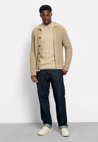 Beiger, gerippter Cardigan über einem cremefarbenen, kabelgestrickten Pullover, kombiniert mit dunklen Jeans und weißen Turnschuhen. Schlichte Knopfdetails und klare Linien.