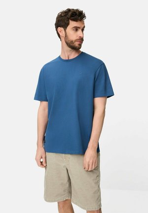 Man met krullend haar en baard, gekleed in een effen blauwe korte mouw t-shirt en beige knielange shorts, kijkt naar links.