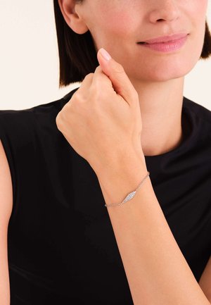 Zilveren armband met een druppelvormige hanger, voorzien van een getextureerd oppervlak en heldere steenaccenten, gedragen om een pols tegen een donkere top.