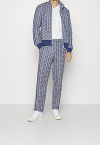 Man med blå mönstrad zipjacka och matchande byxor, bärande vita sneakers och en enkel vit t-shirt, stående mot en vit bakgrund.