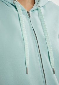 Felpa con zipper azzurro chiaro, caratterizzata da una texture liscia, con lacci decorativi, zip argentata e un design di cappuccio comodo.