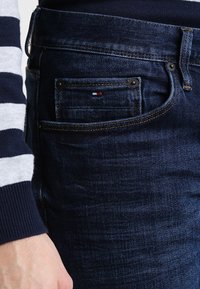 Mörkblå denimjeans med slim fit, som har en liten bakficka prydd med en röd, vit och blå logotyp.