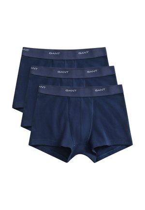 Młody mężczyzna z krótkimi kręconymi włosami, ubrany w granatowe bokserki z logo na pasku, stojący na białym tle.