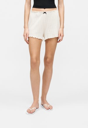 Γυναίκα που φοράει λευκή πλεκτή βερμούδα με βολάν στο τελείωμα και μαύρο φιόγκο, μαύρο κοντό τοπ και λευκές σαγιονάρες, στέκεται σε λευκό φόντο.