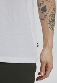 Witte crewneck t-shirt van zachte stof, met een zijsplit en een klein zwart label aan de zoom, gecombineerd met donkergroene broek.