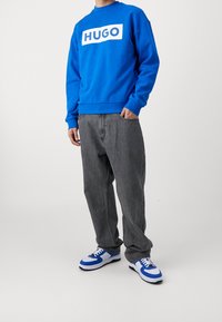 Niebieska bluza z białym logo "HUGO", szare luźne dżinsy oraz niebiesko-białe sneakersy z czarnymi akcentami. Jednolite tło.