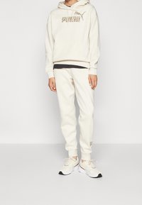 Kremowy hoodie z przednią kieszenią i logo, w połączeniu z pasującymi dresami. Obuwie to lekkie sneakersy w białym i czarnym wzorze.