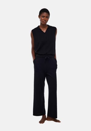 Femme debout pieds nus portant un haut noir sans manches avec col et un pantalon large noir à taille coulissante avec poches.