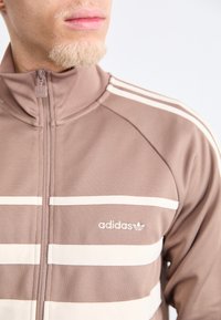 Brown zip-up jacket with white horizontal stripes and logo. Made of soft material, featuring high collar and subtle textured accents.