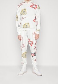 Sudadera blanca con capucha y pantalones de chándal que presentan coloridos estampados gráficos, incluyendo números y anillos de campeonato. Zapatillas blancas completan el conjunto.