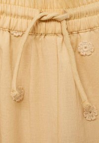 Tissu beige clair avec taille froncée et cordon de serrage. Présente des fleurs brodées décoratives dans une couleur claire contrastante.