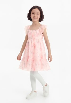 Fille souriante portant une robe fleurie rose avec des manches à volants, des leggings blancs et des baskets blanches, se tenant sur une jambe sur un fond blanc.