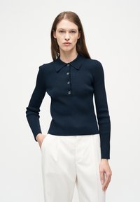 Γυναίκα με μακρυμάνικο μπλε πόλο με ρίγες και τέσσερα κουμπιά, συνδυασμένο με ψηλόμεσο λευκό παντελόνι με πιέτες, στέκεται μπροστά σε απλό φόντο.