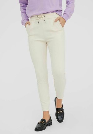 Femme portant un pantalon crème à cordon avec poches latérales et des mocassins noirs, debout les mains dans les poches et un pied en avant.