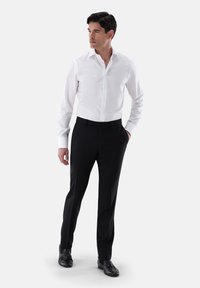 Weißes Hemd mit spitzem Kragen und schmaler Passform, kombiniert mit schwarzen maßgeschneiderten Hosen und schwarzen Schuhen, vor neutralem Hintergrund.