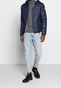 Marinblå vadderad jacka med huva, texturerad yta, grå polotröja, ljusblå jeans och svarta snörstövlar med kraftiga sulor.