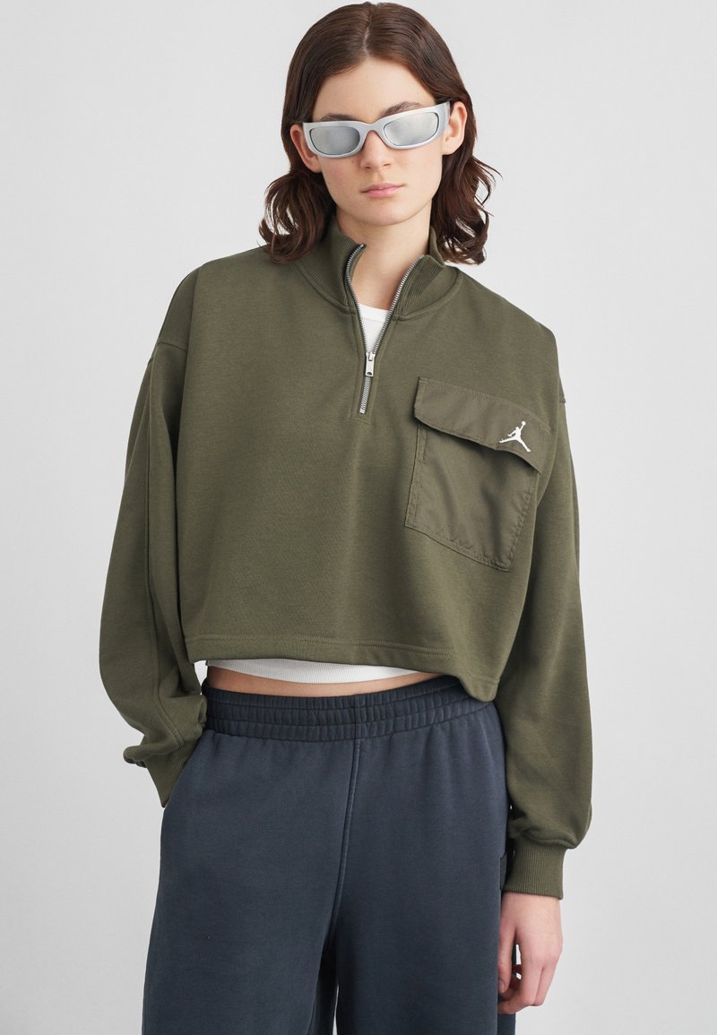 Γυναίκα φορεμένη με ελιά πράσινη αμάνικη ζακέτα με φερμουάρ και τσέπη, ναυτικές μπλε παντελόνες, καθρέφτες ασημένιες γυαλιά ηλίου, πάνω από απλό φόντο.