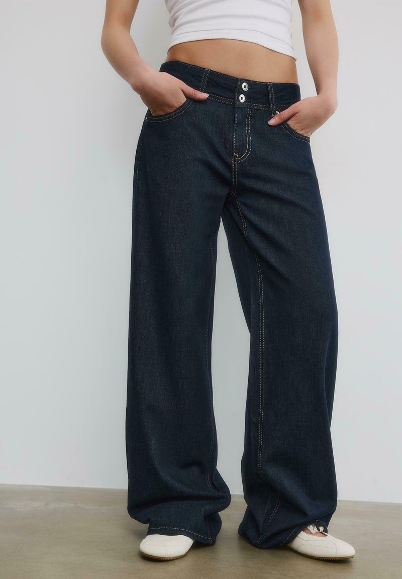 Personne portant un jean large sombre et des chaussures blanches, les mains dans les poches avant, debout sur un sol gris.