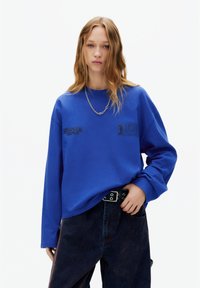 Bluză albastră cu mâneci lungi cu imprimeuri „Stranger Things” și „Classified”. Are croială lejeră, guler rotund ribbed și material texturat.