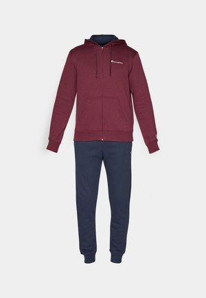 Felpa con zip color bordeaux dotata di cappuccio con cordino, caratterizzata da una tasca frontale e un logo bianco. Abbinata a pantaloni della tuta blu navy, con polsini elasticizzati alle caviglie.