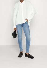 Camisa blanca de botones, vaqueros ajustados azules y mocasines negros. Sosteniendo un pequeño bolso negro. Diseño simple y paleta de colores neutros.