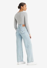 Jean taille haute bleu clair avec une coupe décontractée, présentant des poches arrière et un patch en cuir de la marque. Associé à un haut gris à manches longues.