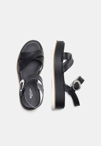 Sandalias de cuero negro con tiras cruzadas, una hebilla redonda y una suela gruesa blanca con un diseño de agarre texturizado.