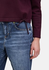 Pull en coton bourgogne avec un détail de lien à l'ourlet, associé à un jean en denim bleu avec une coupe classique et des éléments légèrement décolorés.