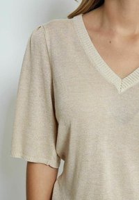 Beige pletený top s výstřihem do V a krátkými rukávy, který má kolem výstřihu žebrovanou texturu a mírně volný střih.