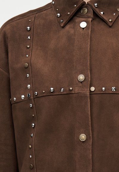 Καφέ τζάκετ από σουέτ με ασημένια και μπρούντζινα μεταλλικά καρφιά τοποθετημένα σε οριζόντια και κάθετα μοτίβα, με κουμπιά τύπου κλιπ και μυτερό γιακά.