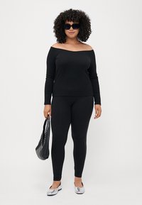 Crna majica s dugim rukavima i off-shoulder izrezom uparena s crnim tajicama. Ima srebrne zakovice sa strane. Model drži crnu torbu i nosi sunčane naočale.