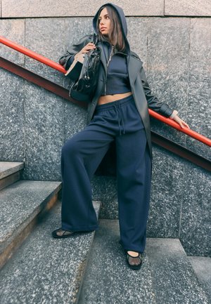 Jeune femme en veste à capuche sombre et pantalon large, debout sur des escaliers en pierre grise, tenant la bandoulière d'un sac noir et appuyée sur une rambarde rouge.