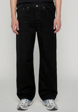 Person iført løstsiddende sorte jeans kombineret med hvide og blå atletiksko stående mod en ensfarvet baggrund.