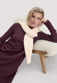 Blonde Frau in einem dunkelpurpurnen Langarmkleid und cremefarbener Schal lehnt an einem cremefarbigen strukturierten Sessel mit Holzbeinen vor einem einfarbigen Hintergrund.