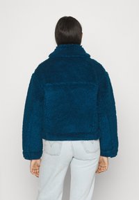 Giacca in pelliccia eco-teal tagliata, con colletto alto e vestibilità oversize. Presenta maniche lunghe e un dettaglio di tasca posteriore visibile.