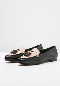 Mocassins en cuir noir avec une empeigne en cuir beige, garnitures dorées et pompons noirs. Design élégant, semelle plate et texture lisse.