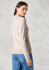 Heller beiger Pullover aus weichem Strickmaterial mit gerippter Textur. Mit langen Ärmeln und einem taillierten unteren Saum. Kombiniert mit blauen Jeans.