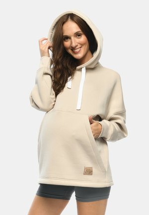 Beige hoodie with white drawstrings, large front pocket, and brown logo patch. Soft fabric, relaxed fit, and oversized hood design.