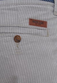 Tissu gris à fines rayures avec un bouton marron et une étiquette en cuir indiquant « INDICODE EST. 1993 » au-dessus d’une fente de poche.