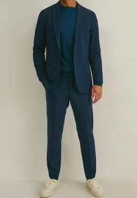 Costume bleu marine avec pantalon assorti, en tissu texturé. Porté sur un pull bleu foncé ; complété par des baskets blanches.
