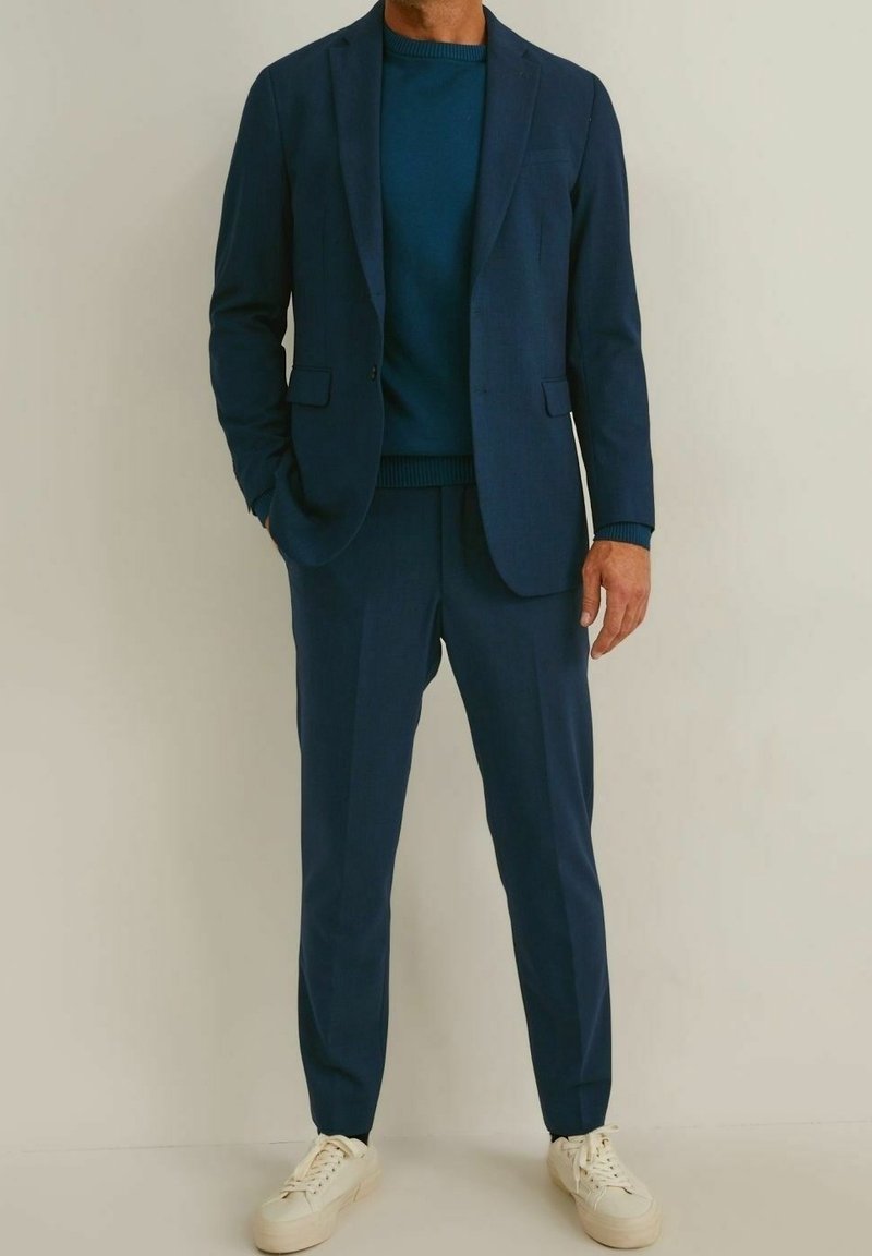Costume bleu marine avec pantalon assorti, en tissu texturé. Porté sur un pull bleu foncé ; complété par des baskets blanches.