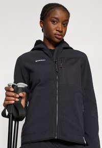 Giacca in pile nera con zip frontale, cappuccio e pannello frontale testurizzato. Presenta un logo, due tasche con zip e una finitura liscia. Mano che afferra dei bastoni da trekking.