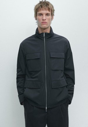 Giovane uomo con capelli chiari che indossa una giacca nera con cerniera e quattro tasche a patta, in piedi davanti a uno sfondo grigio chiaro uniforme.