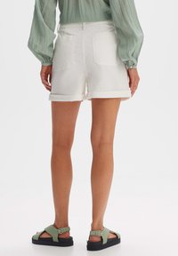 Shorts in denim bianco con risvolti, due tasche posteriori, abbinati a sandali verdi con fibbia. Il tessuto ha una superficie liscia.