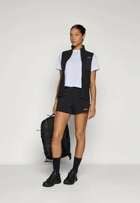 Colete preto sobre uma t-shirt clara, combinado com calções pretos. Uma mochila preta, sapatilhas elegantes e meias escuras completam o visual. Design simples e funcional.